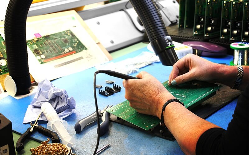 A pair of women's hands working on an electric circuit board with a soldering iron and vacuum hose to remove smoke and dust particles.
